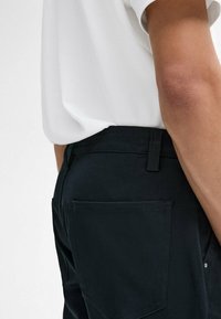Black trousers with a smooth texture, featuring a back pocket and a belt loop. Paired with a plain white t-shirt, taken from a slight side angle.