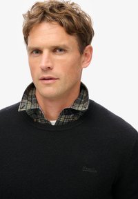 Zwarte trui met een gestructureerde brei, gelaagd over een geruit flanellen shirt. Heeft een ronde hals en een subtiele logo op de borst.