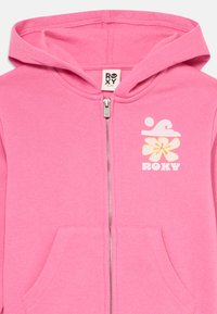 Rosa hettejakke med glidelås, frontlommer og hvit-gul blomst og bølge logo med "ROXY" tekst på brystet.