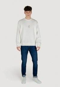 Weißes Sweatshirt mit Rundhalsausschnitt und strukturierter Logo-Applikation auf der Brust, kombiniert mit dunkelblauen Jeans und weißen Sneakern.