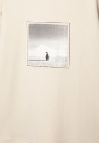 Beige sweatshirt med et firkantet sort-hvidt foto af en pingvin, med tekst nedenunder: "Jeg tror ikke, dette er mit sted."