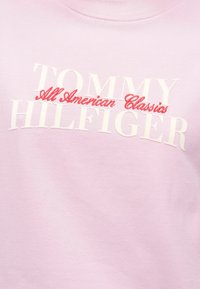 Hellrosa Sweatshirt mit kräftigem weißen und roten Schriftzug "TOMMY HILFIGER" und "All American Classics." Weiches Baumwollmaterial.
