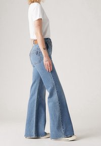 Helle blaue Schlagjeans aus Denim, hoch geschnitten mit einer abgespeckten Waschung, ausgestattet mit Gesäßtaschen und einem getragenen Saum, kombiniert mit einem weißen T-Shirt.