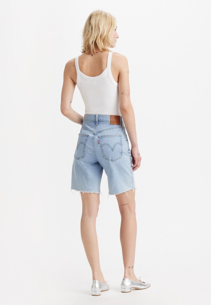 Shorts en denim bleu clair avec un ourlet effiloché, taille mi-haute, et une étiquette en cuir marron à l'arrière de la taille. Associés à un débardeur blanc et des chaussures argentées.