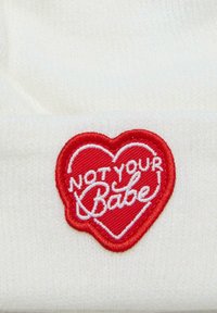 Biely pletený klobúk s červeným srdcovým nášivkom a textom "NOT YOUR Babe" v bielej kurzívnej výšivke.