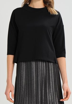 Vrouw die een zwarte top met ellebooglange mouwen draagt, gecombineerd met een metallic plissérok met hoge taille.