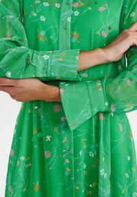 Grön blommig klänning med knäppning framtill, långa ärmar och olika blommönster i rosa, vitt och guld. Genomskinligt tyg.