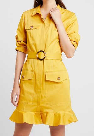 Mujer con vestido amarillo con cinturón, mangas remangadas, bolsillos con solapa y botones, y bajo con volantes, de pie frente a un fondo liso.