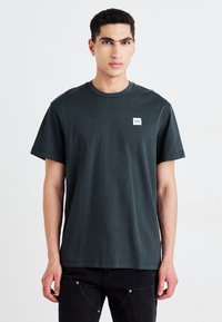 T-shirt in cotone verde scuro con maniche corte, collo rotondo e un piccolo etichetta bianca sul petto. Presenta una vestibilità rilassata.