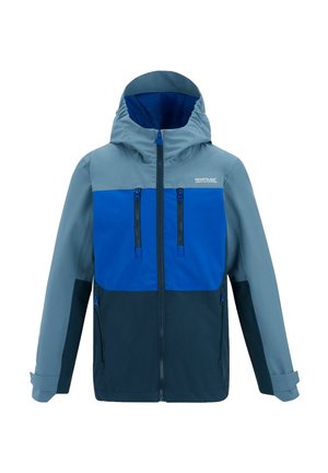 Garçon portant une veste bleue et grise, un pantalon bleu marine et des chaussures de randonnée, debout sur un fond clair uni.