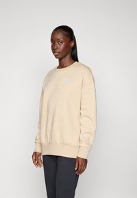 Kvinne med mørk hud og langt svart hår iført en beige Under Armour hettegenser og svarte bukser, står foran en lys, ensfarget bakgrunn.