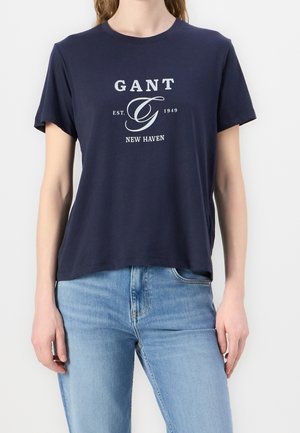 Personne portant un T-shirt GANT bleu marine avec un logo blanc "GANT EST. 1949 NEW HAVEN" et un jean bleu clair, debout devant un fond blanc.