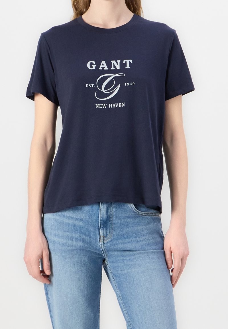Henkilö pukeutunut tummansiniseen GANT T-paitaan, jossa valkoinen logo "GANT EST. 1949 NEW HAVEN", ja vaaleansinisiin farkkuihin, seisoo valkoista taustaa vasten.