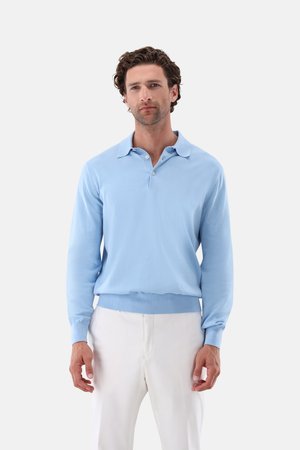 Mann mit lockigem Haar, der ein hellblaues Langarm-Poloshirt und weiße Hose trägt, vor einem schlichten weißen Hintergrund stehend.