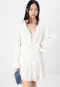 Λευκό φόρεμα με ρυάκι στον λαιμό και τις μανσέτες, υφαντό ύφασμα και κλείσιμο με κορδόνι. Μοντέλο κρατάει μια μικρή μπλε τσάντα.