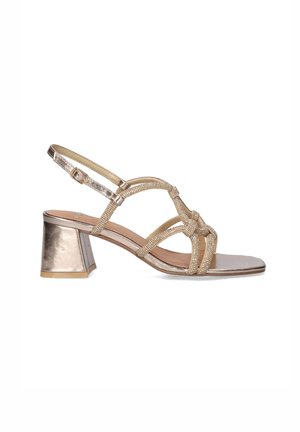 Sandalia dorada con tiras, tacón grueso, tiras delanteras anudadas y hebilla ajustable en el tobillo, mostrada de perfil sobre fondo blanco.