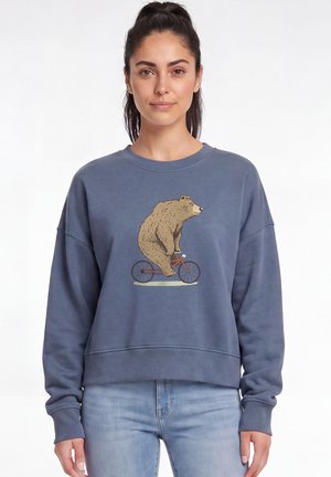 Femme portant un sweat-shirt bleu avec un motif d’un ours à vélo rouge, associé à un jean bleu clair, sur un fond blanc uni.