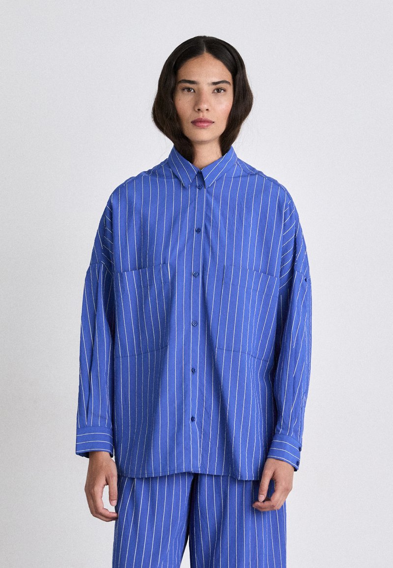Camicia a bottone a righe blu con colletto, dotata di due tasche sul petto, maniche lunghe e una vestibilità morbida. Tessuto in cotone liscio.
