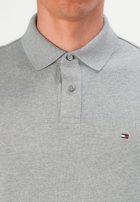 Mężczyzna w szarej koszulce polo z dwiema guzikami przy dekolcie oraz małym haftowanym logo w kolorach czerwonym, białym i niebieskim na piersi.