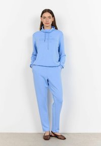 Lichtblauwe sweatshirt met een hoge kraag, trekkoord en bedrukte tekst. Bijpassende losvallende broek in dezelfde kleur. Gecombineerd met platte schoenen.