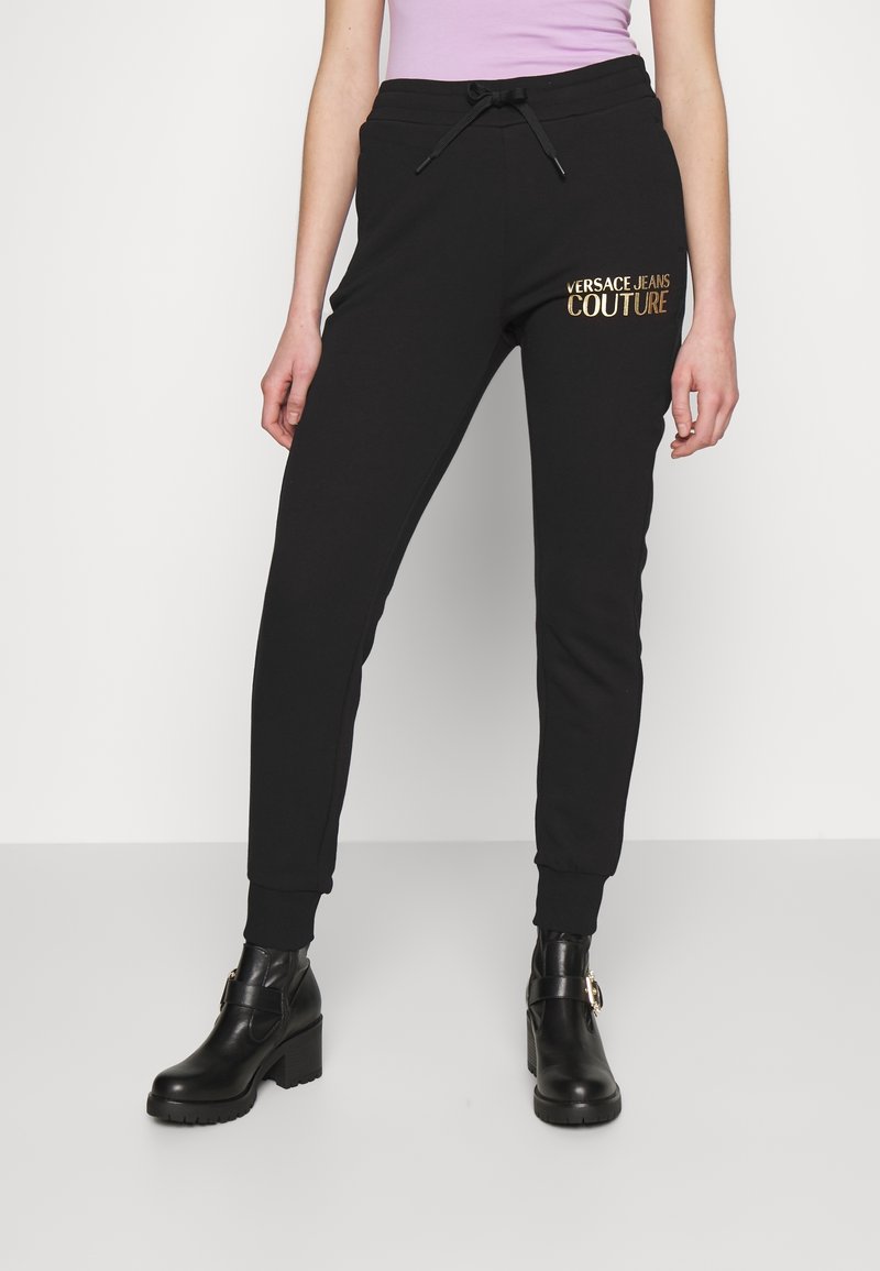 Femme portant un pantalon de jogging noir Versace Jeans Couture à taille coulissante et des bottines noires à talons.
