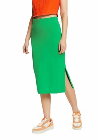 Jupe midi verte avec une fente latérale, en tissu lisse. Associée à des baskets orange et blanches avec des accents contrastants.