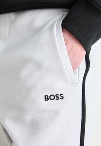 Hellgraue Sweatpants mit elastischem Bund, seitlicher Tasche und einem schwarzen Logo. Der Stoff hat eine glatte Textur mit kontrastierenden schwarzen Akzenten.