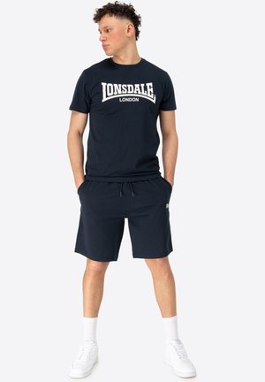 Mornarsko plava pamučna majica s bijelim grafičkim natpisom "Lonsdale London", u kombinaciji s mornarsko plavim kratkim hlačama. Model nosi bijele tenisice i bijele čarape do koljena.