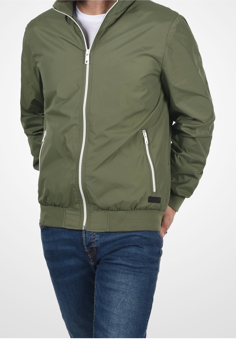 Homme portant une veste zippée vert olive avec une fermeture éclair blanche et un jean bleu, les mains dans les poches de la veste, sur un fond uni.