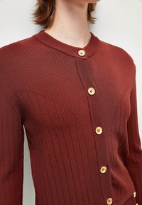 Rdzawy, prążkowany kardigan z okrągłym dekoltem, ozdobiony sześcioma drewnianymi guzikami, z kontrastującą fakturą i subtelnymi szwami dla dodatkowego efektu.
