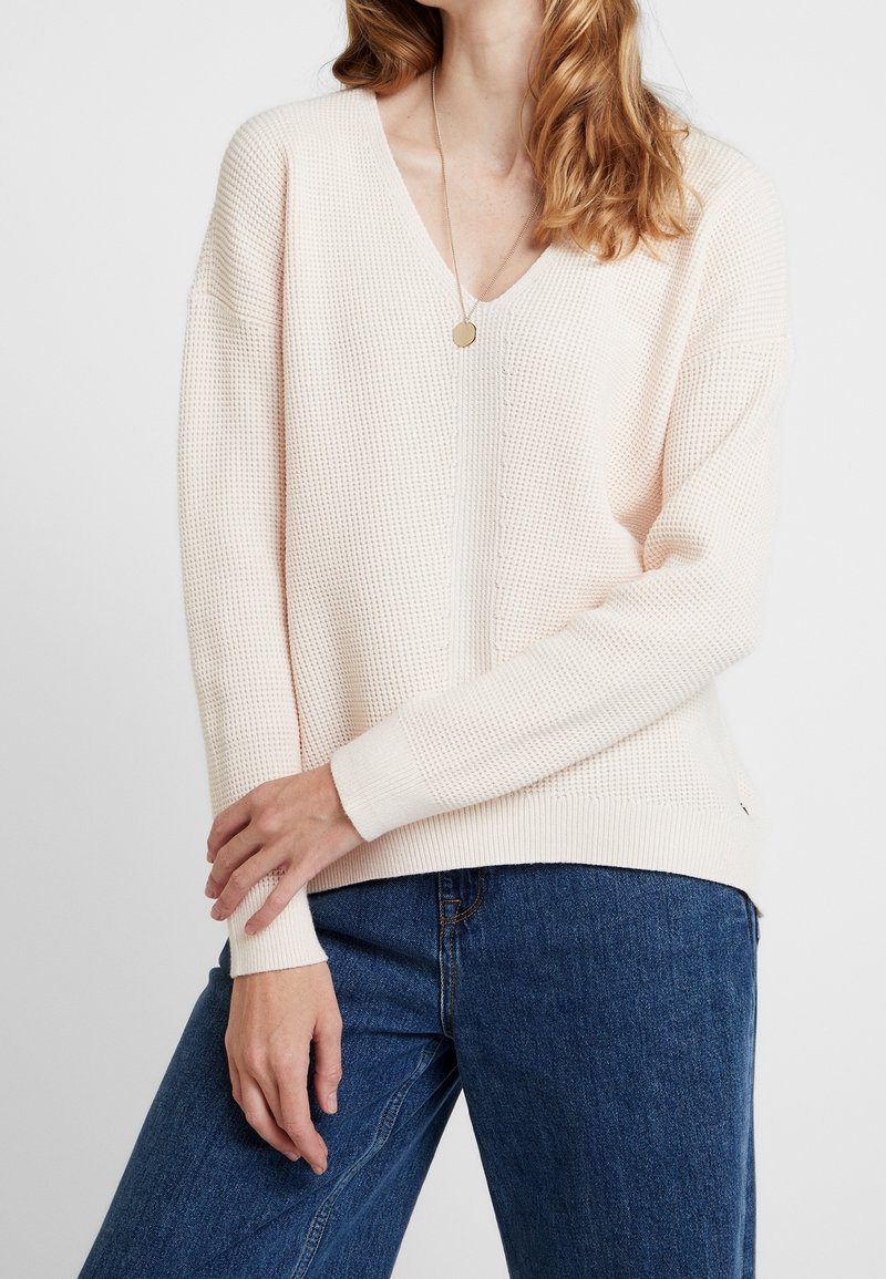 Lichtroze gebreide trui met V-hals, lange mouwen, geribde manchetten en een gestructureerde stof, gecombineerd met donkerblauwe spijkerbroek.