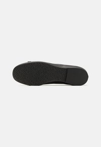 Semelle de chaussure noire avec un motif de prise texturé comportant des lettres en relief sur une base plate à talon bas, sur fond blanc.