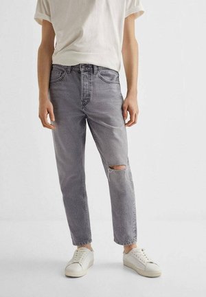 Personne portant un jean déchiré gris clair, un t-shirt blanc et des baskets blanches sur un fond clair uni.