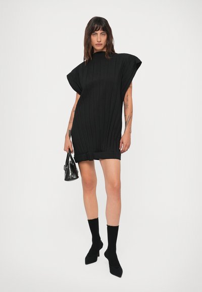 Mulher com cabelo até os ombros, vestindo um vestido mini de malha preta sem mangas, botas de cano curto pretas, segurando uma pequena bolsa preta.