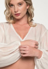 Weiße, halbtransparente Bluse mit V-Ausschnitt, langen Puffärmeln und gerafften Manschetten. Besitzt ein strukturiertes, gestreiftes Muster.