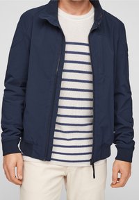 Veste légère bleu marine en tissu texturé, col montant et fermeture éclair, portée sur un pull crème à rayures.