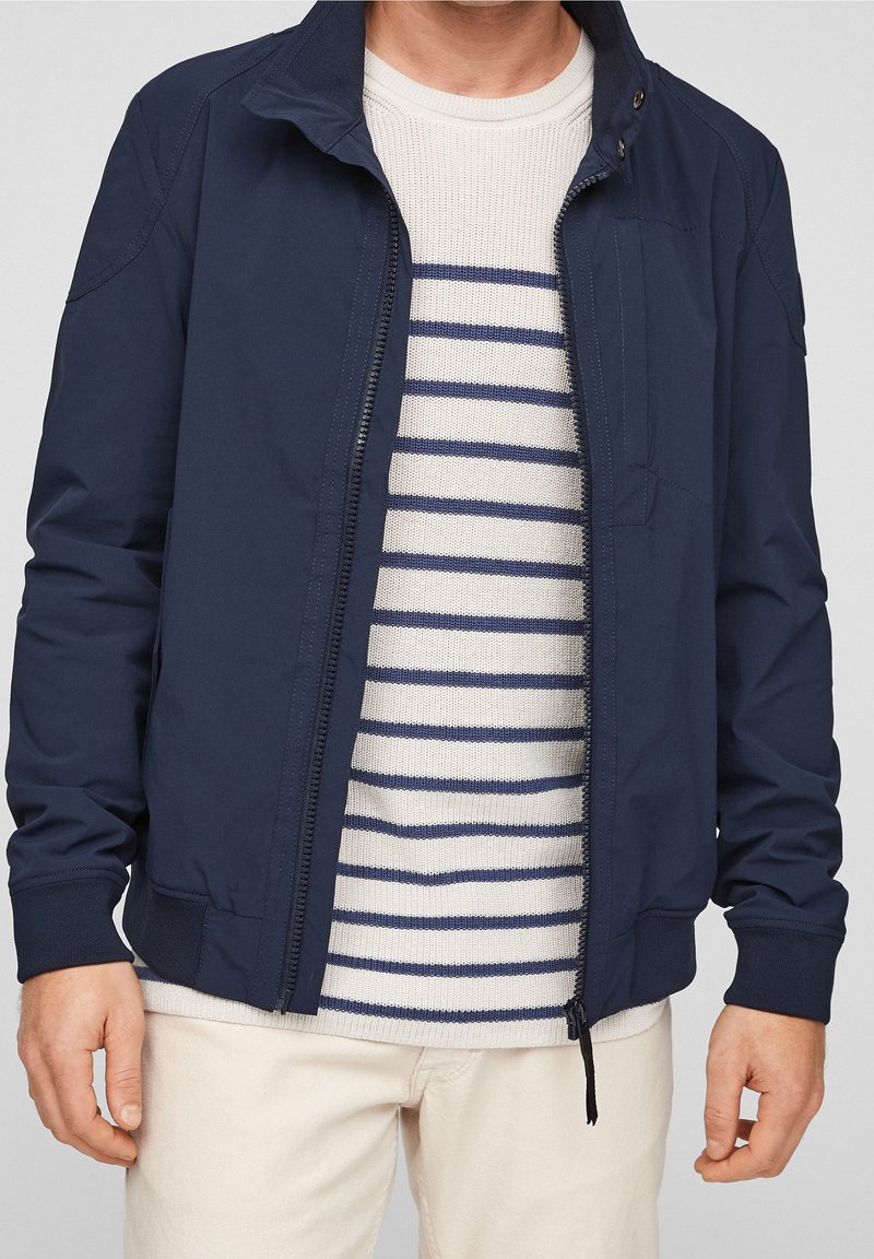 Veste légère bleu marine en tissu texturé, col montant et fermeture éclair, portée sur un pull crème à rayures.