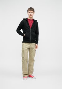 Felpa con cappuccio nera con zip, t-shirt rossa, pantaloni cargo beige, sneaker rosse. Design semplice con tasche sui pantaloni, trame di tessuto lisce.