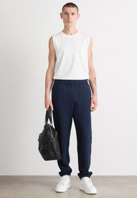 Top sin mangas blanco, pantalones de chándal azul marino con banda elástica en la cintura y zapatillas blancas. Modelo sosteniendo un bolso de cuero negro, de pie contra un fondo neutro.