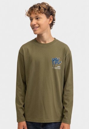 Jeune personne aux cheveux courts et bouclés portant une chemise à manches longues olive avec un motif bleu sur la poitrine, souriante et regardant sur le côté.