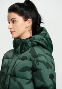 Chaqueta acolchada de camuflaje verde con capucha, que presenta un diseño acolchado y un parche de logo redondo en la manga. Acabado texturizado y brillante.