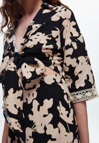 Blusa nera e beige con motivo floreale, davanti con lacci, maniche a tre quarti e bordino in pizzo. Tessuto leggero.