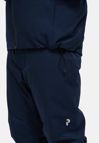 Námořnickomodrá sportovní bunda a kalhoty vyrobené z hladkého materiálu. Bunda má přední zip a nastavitelnou šňůrku v pase.