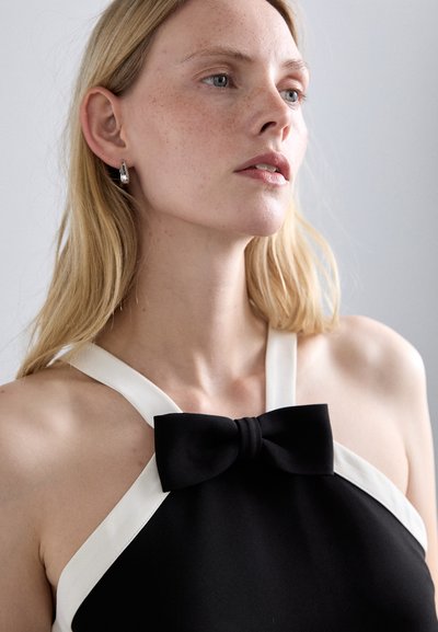 Svart halterneck-top med hvite stropper og en sentral sløyfedetalj. Glatt tekstur med en tettsittende design. Enkel og moderne eleganse.