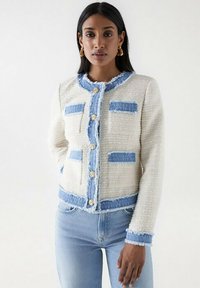 Chaqueta de tweed blanca con acentos de vaquero azul deshilachado, con botones dorados y cuatro bolsillos. Diseño estructurado con cuello redondo.