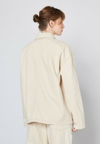 Beige, oversized denimjakke med en glat tekstur, spids krave og syede detaljer, vist fra bagsiden.