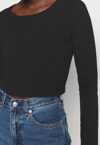 Svart långärmad cropped topp med rund halsringning, ihopparit med högmidjade blå denimjeans som har knappstängning och synlig sömnad.