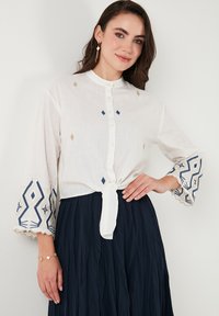 Blusa blanca de botones con lazo delantero, que presenta bordados geométricos en azul y dorado en las mangas acampanadas. Tela ligera y texturizada.