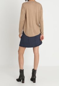 Haut à manches longues marron clair avec un ourlet incurvé, associé à une jupe bleu foncé. Le mannequin porte des bottines grises à talons hauts. Texture de tissu lisse.