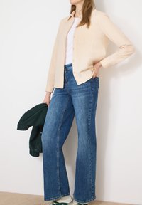 Femme en chemise beige à boutons, t-shirt blanc, jean bleu et baskets vertes tenant une veste vert foncé contre un mur blanc.